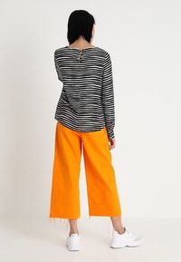 Osoba otočená zády, oblečená v černobílém pruhovaném tričku s dlouhým rukávem, jasně oranžových širokých kalhotách do půli lýtek a bílých teniskách.