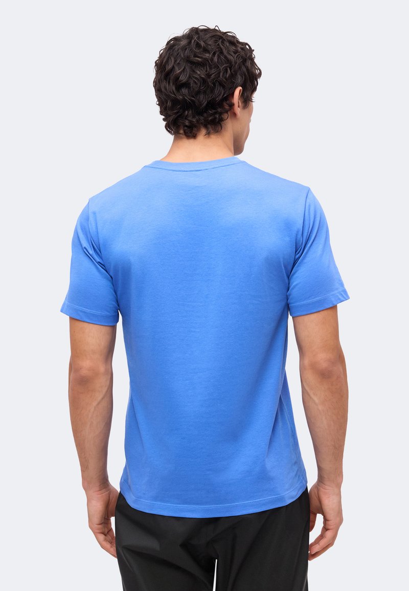 Hombre con cabello rizado y oscuro, vistiendo una camiseta azul lisa de manga corta y pantalones negros, visto desde atrás sobre un fondo claro.