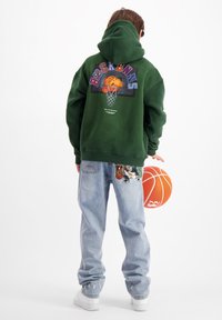 Groene hoodie met grafisch ontwerp van een basketbal en ring. Lichtblauwe jeans met basketbal-thema borduurwerk. Witte sneakers.