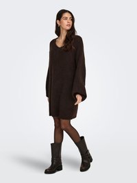 Brown knitted sweater dress with a V-neck and long, wide sleeves, paired with black knee-high boots featuring buckled accents.
