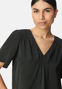 Schwarze Bluse mit V-Ausschnitt, kurzen Puffärmeln und Rüschen an den Schultern. Hergestellt aus einem leichten, glatten Stoff.