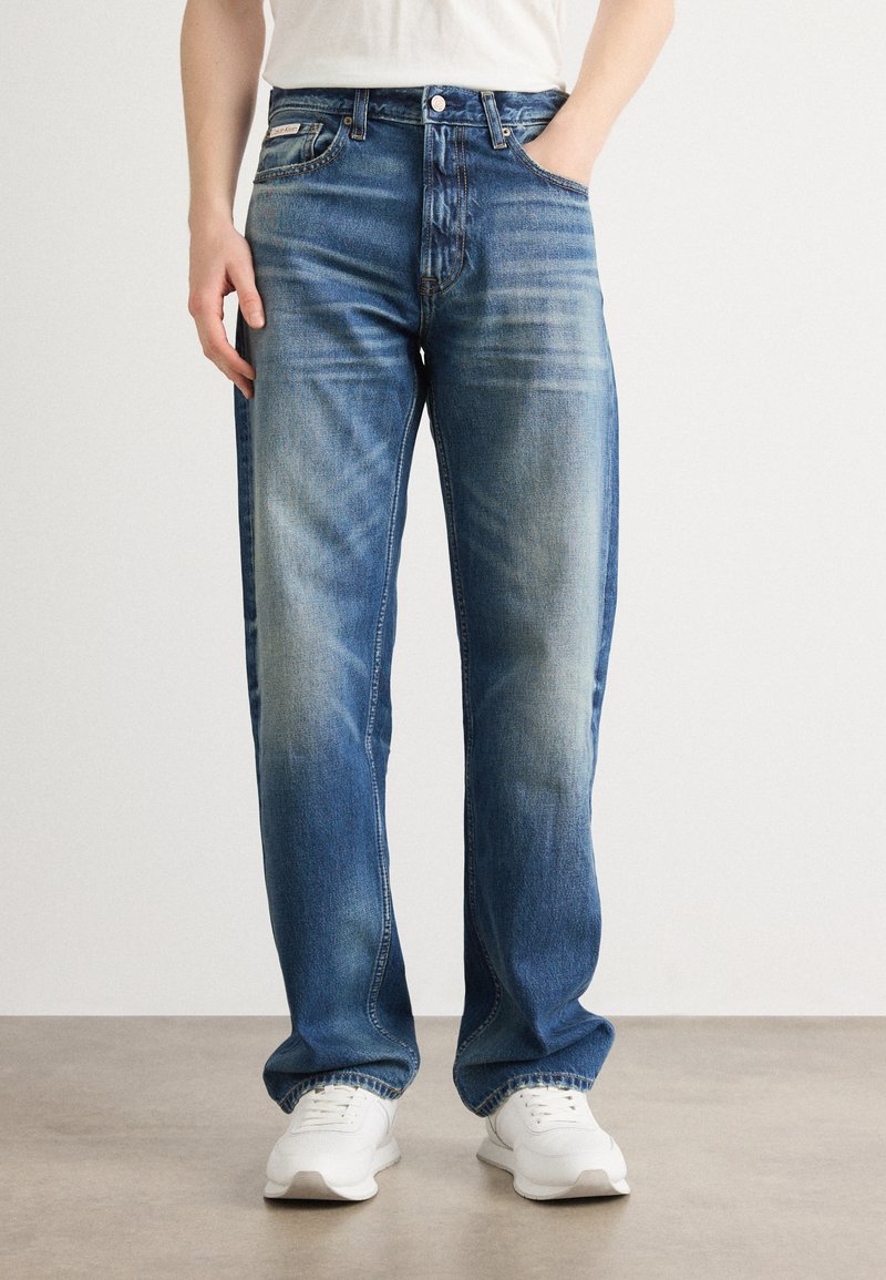 Modri denim traper hlače s ravnim krojem, ki vsebujejo detajle blatenja in obliko s petimi žepi. Uparjene z belimi supergami za kontrast.