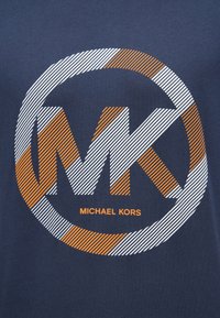 Jūros mėlynos spalvos marškiniai su apvaliu grafiniu dizainu, kuris turi baltą ir oranžinę dryžuotą „MK“ logotipą ir „MICHAEL KORS“ tekstą ryškiai paryškinta šriftu.