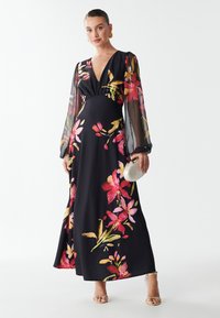 Abito lungo nero con stampa floreale colorata. Presenta un profondo scollo a V e maniche lunghe trasparenti. Tessuto morbido con silhouette fluida, abbinato a una pochette.