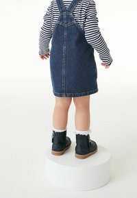 Kleinkind steht auf einer runden weißen Plattform und trägt ein gestreiftes Langarmshirt, ein Jeans-Pinanoerkleid, weiße Rüschenstrümpfe und dunkle Knöchelstiefel.