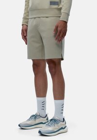 Shorts en coton beige clair avec poches latérales, présentant un logo subtil et des détails de couture. Associés à des baskets claires et des chaussettes blanches.