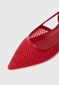Chaussure rouge à bout pointu avec un dessus en maille à motif diamante, accents en vernis rouge brillant, et détails découpés sur les côtés.
