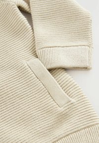 Beige adīts džemperis ar ribotu tekstūru, izteiktām plecu šuvēm un sānu kabatas detaļu. Sastāvā ir mīksta, elastīga dzija, kas nodrošina komfortu.