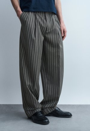 Osoba ubrana w luźne, pionowo paskowane szaro-czarne spodnie oraz czarne eleganckie buty, stojąca na tle jasnoszarego, jednolitego tła.