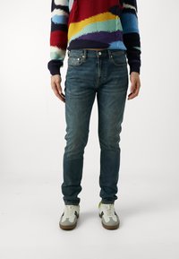 Slim-fit blå denimjeans med en något bleknad finish, ihop med en flerfärgad stickad tröja och vita sneakers med grå detaljer.