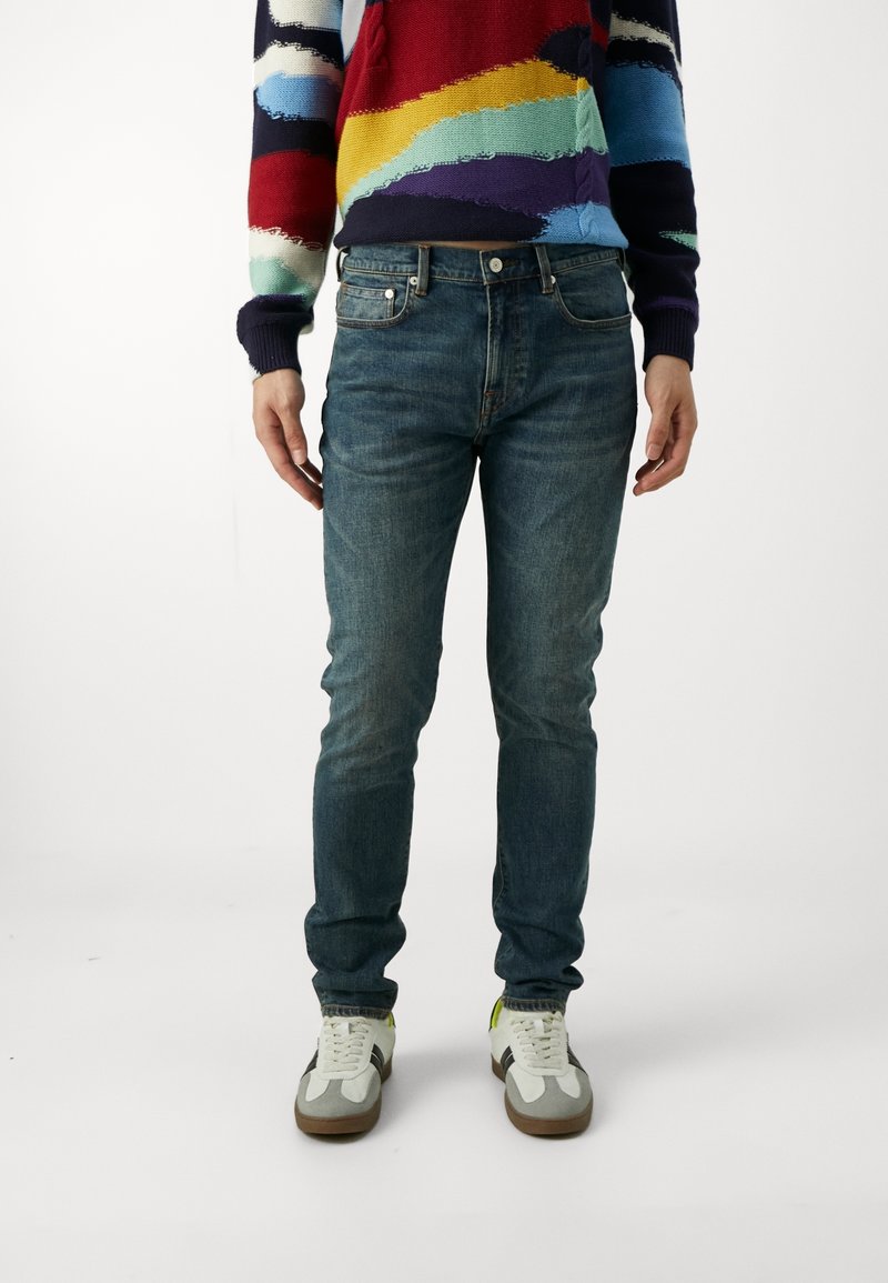 Slim-fit blå denimjeans med en något bleknad finish, ihop med en flerfärgad stickad tröja och vita sneakers med grå detaljer.
