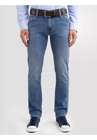 Mees sinistes teksapükstes, pruuni nahkvöö, helesinise särgi ja tumesiniste tennistega, seisab ühevärvilise tausta ees.