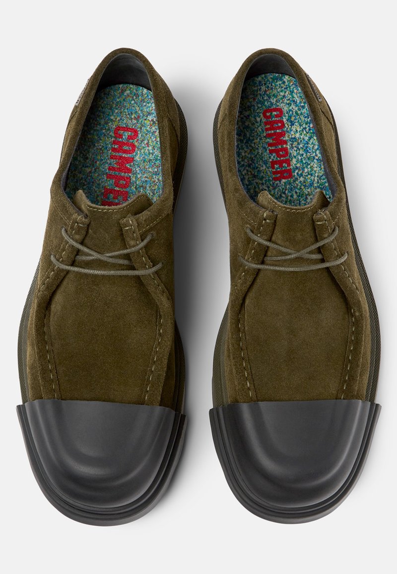 Olivgrüne Wildlederschuhe mit schwarzen Gummikappen an den Zehen und grauen Schnürsenkeln. Bunt gesprenkelte Innensohlen mit "CAMPER" in Rot.