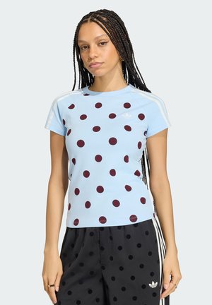 Jonge vrouw met lange vlechten, gekleed in een lichtblauw Adidas T-shirt met polkadots en zwarte polkadotbroek met witte strepen, staand tegen een effen achtergrond.