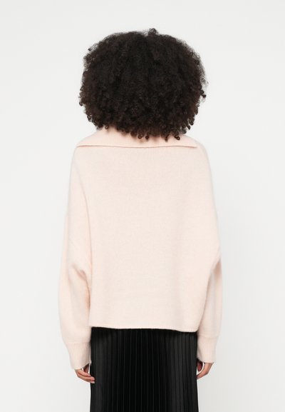 Pull oversize rose pâle avec un large col, présentant une finition en tricot texturé. Stylé avec une jupe plissée noire, mettant en valeur un design superposé.