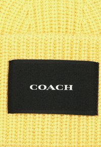Gorro de punto amarillo con una etiqueta rectangular negra que muestra "COACH" en letras blancas. Textura suave, diseño acanalado.