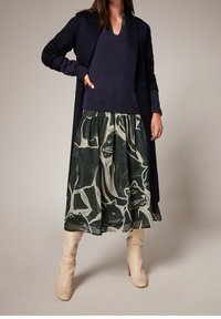 Vrouw in een marineblauwe trui en jas, een groene rok met grote zwarte kattenprints, en off-white kniehoge laarzen, staand met één hand op haar heup.