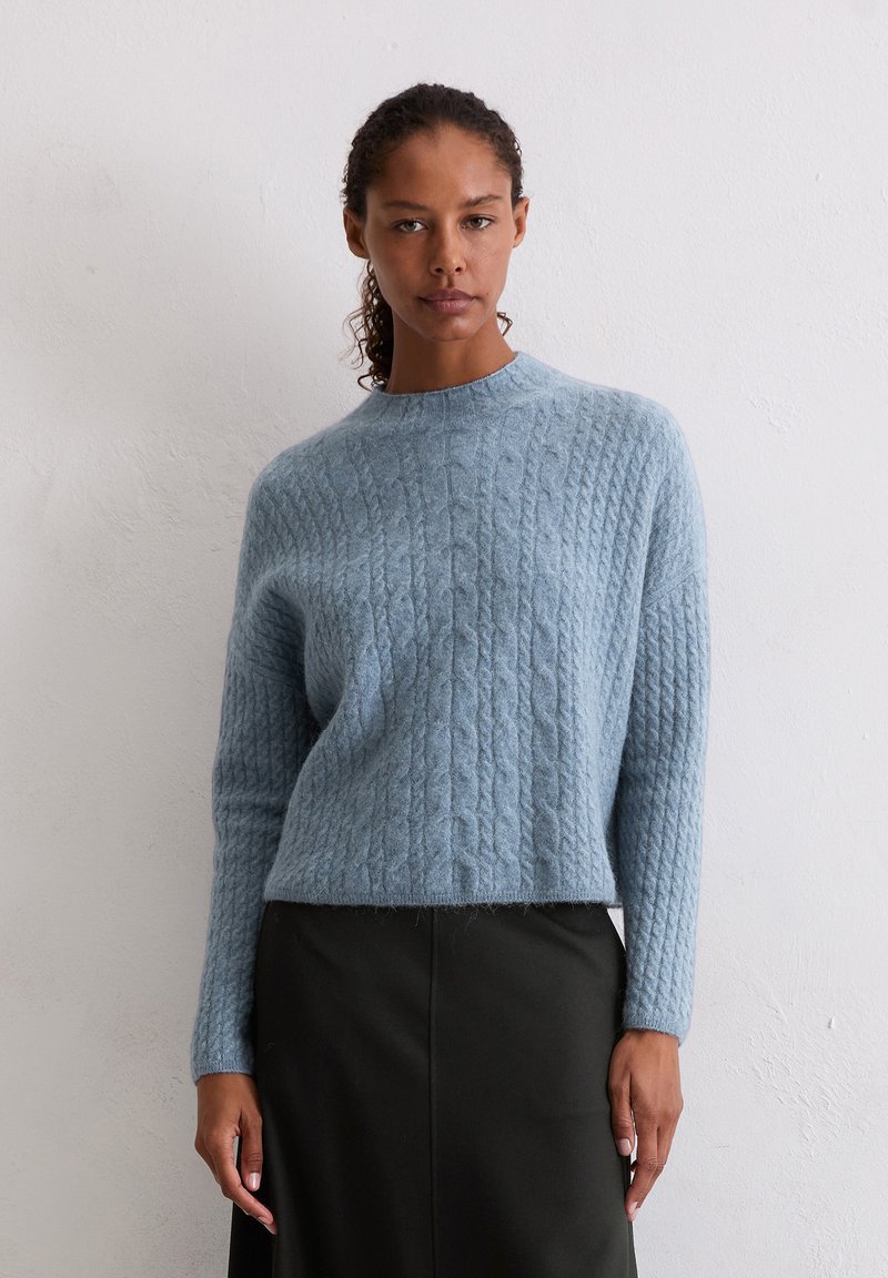 Hellblauer Strickpullover mit einem strukturierten Zopfmuster und einem rundem Halsausschnitt. Kombiniert mit einem schwarzen Midikleid und halblangen Ärmeln.