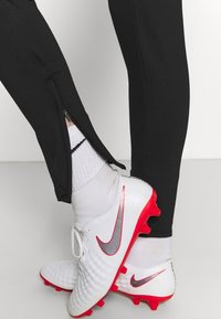 Svarta idrottsbyxor med dragkedjor i nederkant, tillsammans med vita och röda fotbollsskor med en silverfärgad swoosh och texturerad ovandel. Vita strumpor synliga.