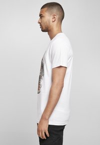 Weißes Baumwoll-T-Shirt mit einem grafischen Druck auf der Vorderseite, kurzen Ärmeln und entspannter Passform. Modell seitlich betrachtet.
