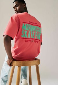 Koral oversized t-shirt med grøn tekst, der læser "MORNINGSIDE SKATE PARK" samt stedoplysninger. Model siddende på en træstol.