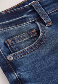 Primo piano di jeans di denim che mostra la tasca frontale, la piccola tasca porta monete, il passante per la cintura e dettagli di cuciture arancioni su tessuto blu.