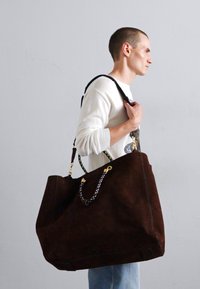 Geantă tote din suede maro cu mânere împletite, accente din aur și un design spațios, prezentată fiind purtată de o persoană.