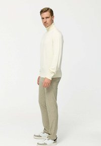 Tejmeszes krémszínű garbó, texturált. Szürke mintás nadrág. Fehér sportcipő szürke részletekkel. A modell oldalra fordulva pózol egy egyszerű háttér előtt.