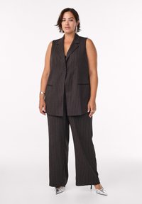 Tumma pinstripes-puku, jossa on hihaton bleiseri ja leveälahkeiset housut. Korostettu hopeanvärisillä korkokengillä ja metallijalokivillä.