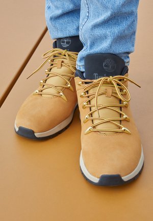 Tanfarvede læderruteboots med gule snørebånd båret med blå jeans på en tan overflade, der viser mærkelogo på den polstrede tung.