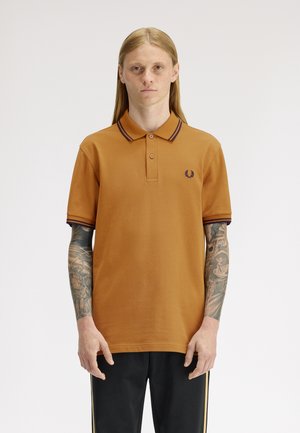 Ung person med langt hår iført brun poloshirt og sorte bukser, med fuldlængde tatoveringer på begge arme, stående mod en hvid baggrund.