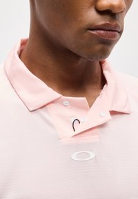 Κοντινό πλάνο ενός ατόμου που φοράει ανοιχτό ροζ πόλο μπλούζα με τρία λευκά κουμπιά και ένα λευκό οβάλ λογότυπο στο στήθος.