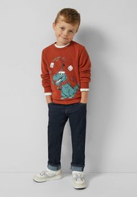 Rode sweatshirt met een blauwe dinosaurusafbeelding en tekst, gecombineerd met donkere spijkerbroek en witte sneakers met grijze accenten.