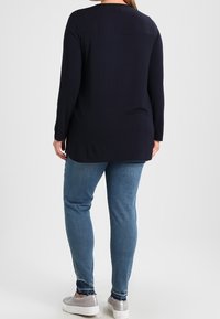 Dlouhý rukáv, tmavě modrý top s kulatým výstřihem a volným střihem, kombinovaný se světle modrými skinny džínami a šedými nazouvacími botami.