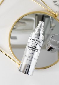 Sølv sprayflaske med pumpe, mærket "Intensiv strækmærke induktions terapi gel," placeret på et rundt spejl med guldkant. 80 ml.