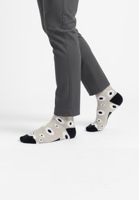 Pies con calcetines beige claro con dedos y talón negros y patrones de ojos en blanco y negro, combinados con pantalones gris oscuro.