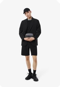 Blazer nero sopra una t-shirt nera, pantaloni corti abbinati e sneakers nere. Tiene un pochette con motivo geometrico e accenti gialli.