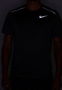 Černé tričko Nike s reflexními akcenty na ramenou, krátkými rukávy a texturovanou tkaninou. Nosené s vzorovanými černými šortkami.
