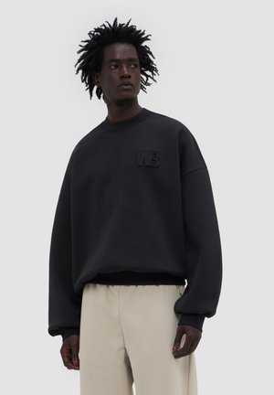 Mężczyzna o teksturowanych włosach nosi oversize czarną bluzę z logo "WB", w połączeniu z jasnobeżowymi spodniami, stojąc na jednolitym tle.