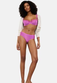 Sieviete ar čirkainiem matiem, ģērbusies gaiši violetā mežģīņu krūšturī un atbilstošos apakšbiksēs, ar baltu kardiganu apsedzot plecus.