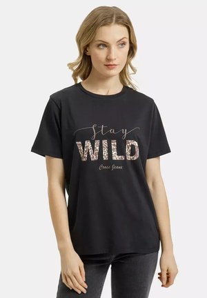 Młoda kobieta ubrana w czarną koszulkę z napisem "stay WILD Cross Jeans", w ciemnych jeansach, stojąca na białym tle.