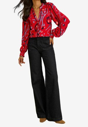 Kobieta w czerwonej bluzce w wzory z bufkami na rękawach oraz w czarnych, szerokich, wysokich dżinsach z brązowymi, spiczastymi butami.