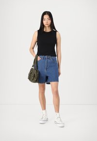 Black sleeveless top tucked into high-waisted light blue denim shorts. Model carries an olive green bag and wears white high-top sneakers.