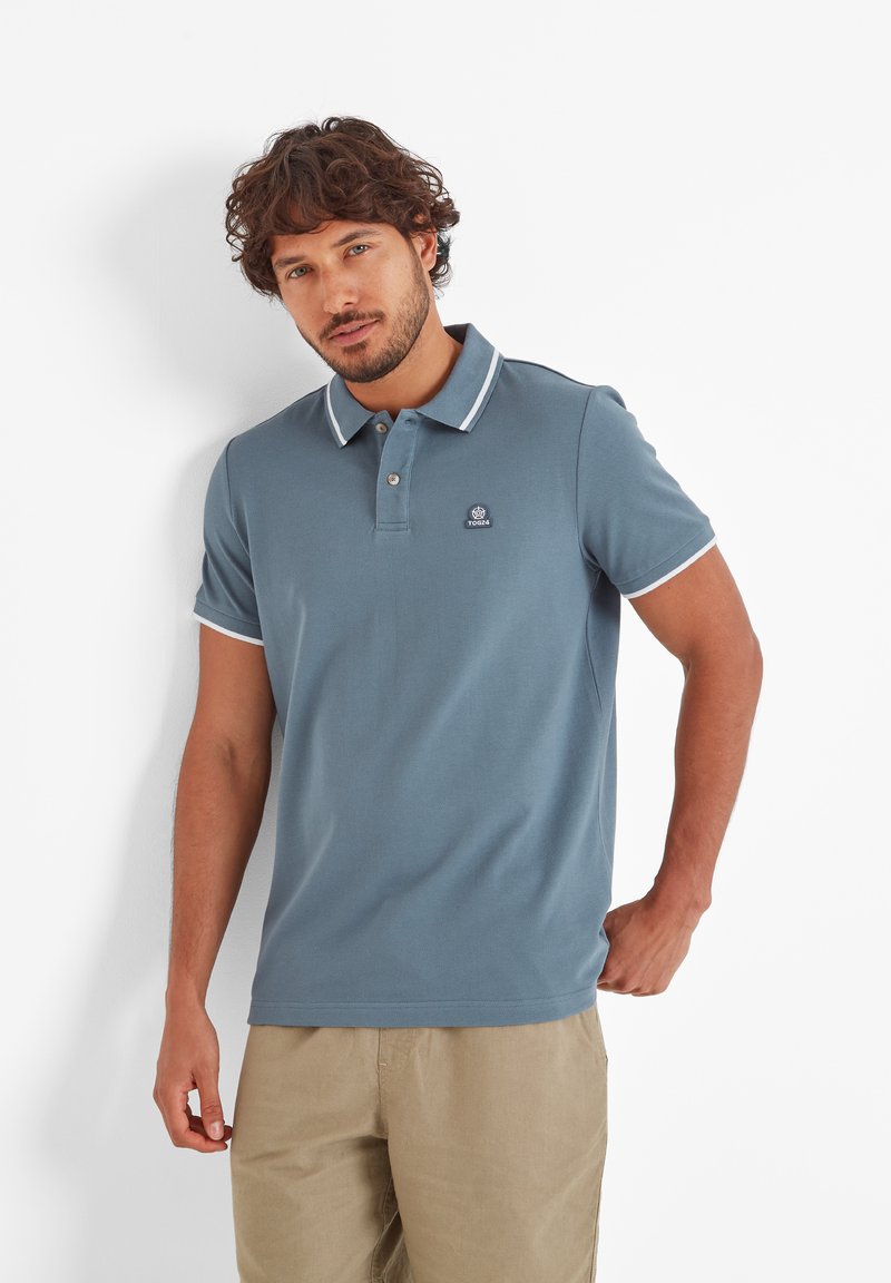 Man with curly hair wearing a blue polo shirt with white trim and beige pants, standing against a white background.