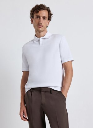 Mann mit lockigem Haar, der ein weißes kurzärmeliges Poloshirt und dunkelbraune Hose trägt, steht vor einem schlichten hellen Hintergrund.