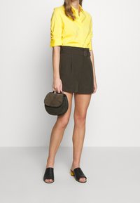 Gul knäpp skjorta med uppvikta ärmar, mörkgröna högmidjade shorts med bälte, svarta slidesandaler och en liten rund handväska.