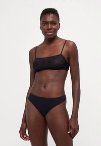 Ženska s kratkimi lasmi nosi črn modrček na naramnice in usklajeno črno spodnje perilo, stoji ob enobarvni svetli ozadju.