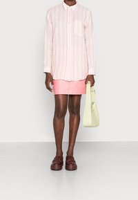 Camicia a righe rosa chiaro con bottoni, gonna mini in pelle rosa, borsa verde oliva e scarpe slip-on bordeaux indossate da un modello contro uno sfondo neutro.