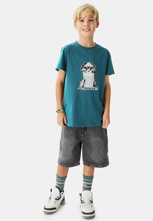 Blond boy wearing teal graphic t-shirt, gray knee-length shorts, striped green socks, white sneakers, and bracelets, standing and smiling.