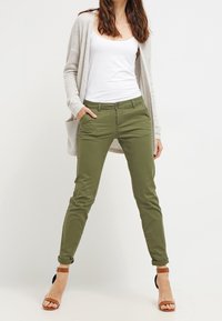 Olive green chinos with a slim fit, featuring a button closure and rolled cuffs, paired with a white tank top and beige cardigan.